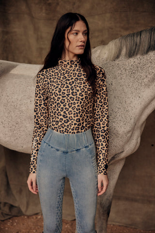 Lifestyle shot of a model in light blue jeans breeches and a leopard print top standing with a grey horse.