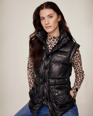 Sitting model wearing the black semi-glossy puffer vest, zipped to show the functional two-way zipper, styled with a leo top and denim breeches.