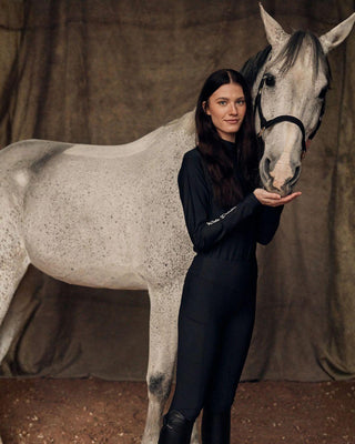 Equestrian lifestyle shot of a model in the all-black riding set cuddling with a grey horse.