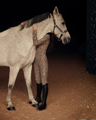 Emotional lifestyle shot of a model wearing the full leopard riding set hugging a grey horse.