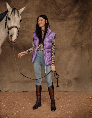 Model in light blue jeans breeches, leo top, and lilac puffer vest holding a grey horse by the lead rope.