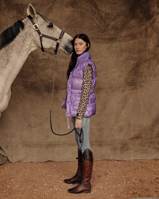 Lifestyle shot of a model in a lilac puffer vest, leo top, and OG denim breeches, standing with a grey horse.