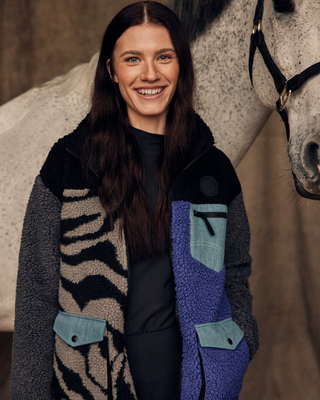 Smiling model in a stable setting wearing an unzipped lilac and zebra teddy jacket with a grey horse in the background.