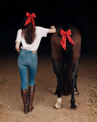 Model from behind in jeans breeches and white t-shirt with a red hair bow, next to a brown horse with a matching red bow.