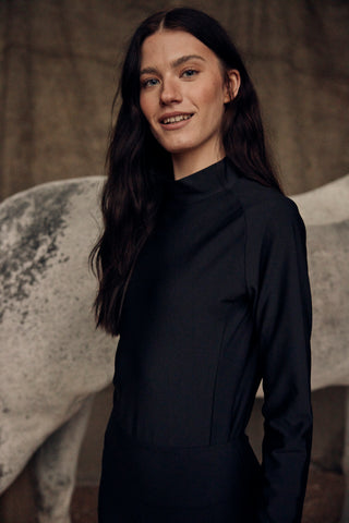 Equestrian portrait of a model wearing the black high-collar riding top with a grey horse in the background.
