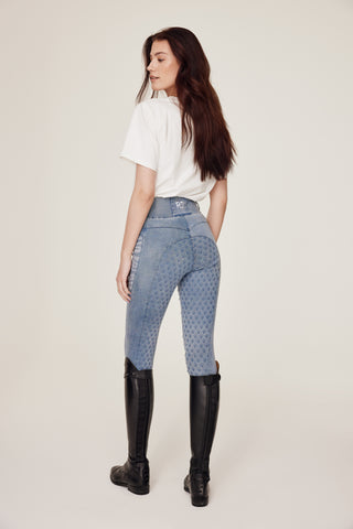 Full body back view of a model in light blue denim breeches and a white horse print t-shirt looking over her shoulder.